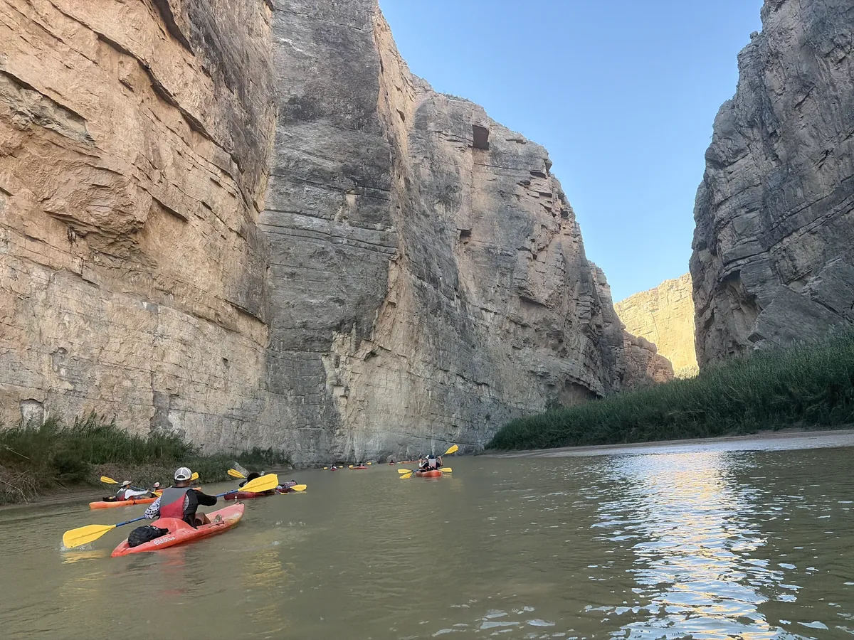 Kayaks moving along a river between tall, sunlit canyon walls.