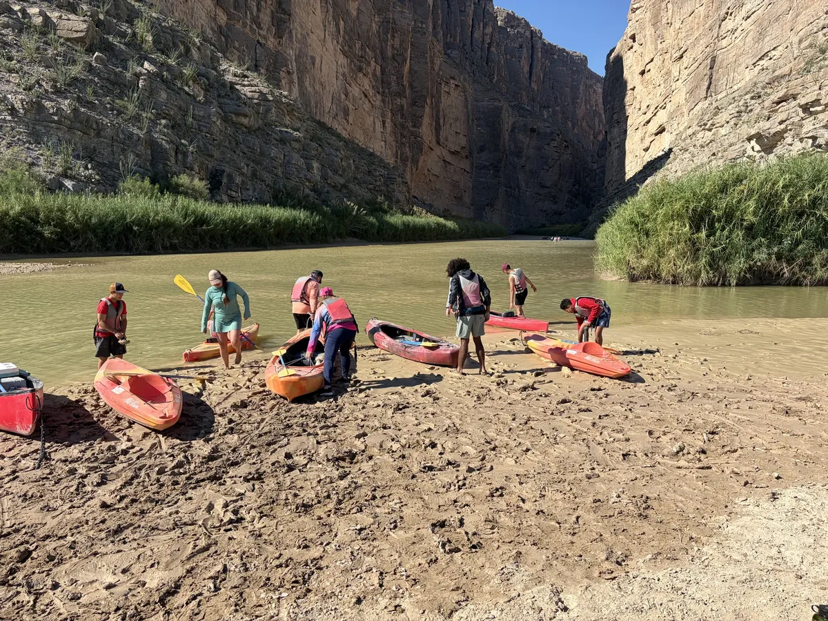 Participants wearing flotation vests prepare colorful kayaks on a muddy riverbank beneath a canyon.