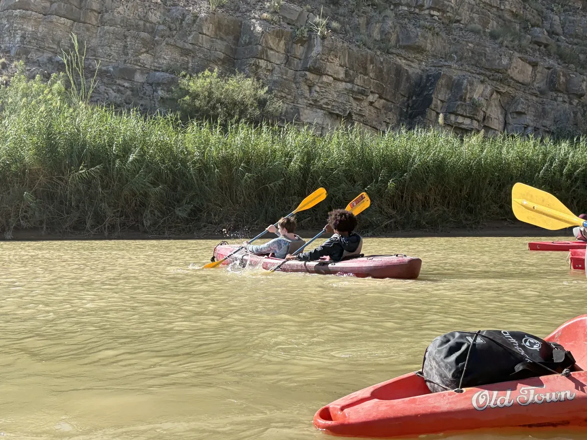 Two paddlers in a kayak raise yellow paddle blades above the river beside green reeds.