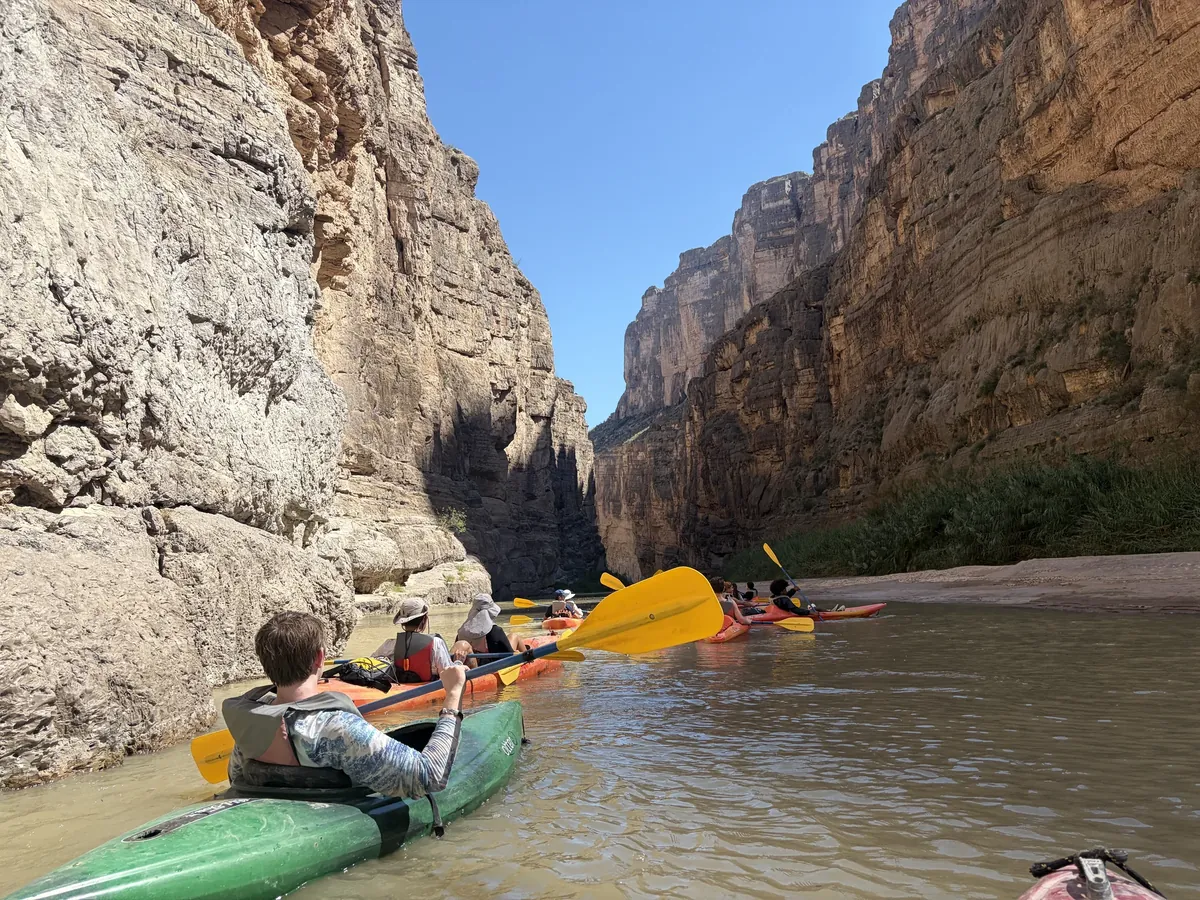 A paddler in a green kayak follows other kayaks between steep canyon walls.