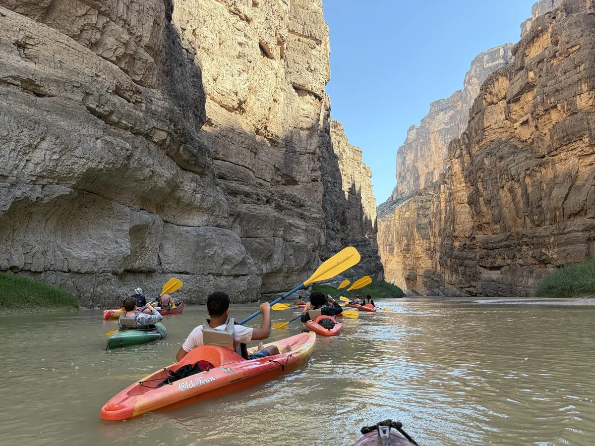 Kayakers with yellow paddles move toward a bright opening between high rock walls.