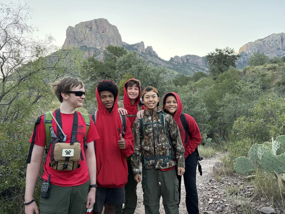 Five Scouts with backpacks standing close together on a trail below rocky mountain ridges.